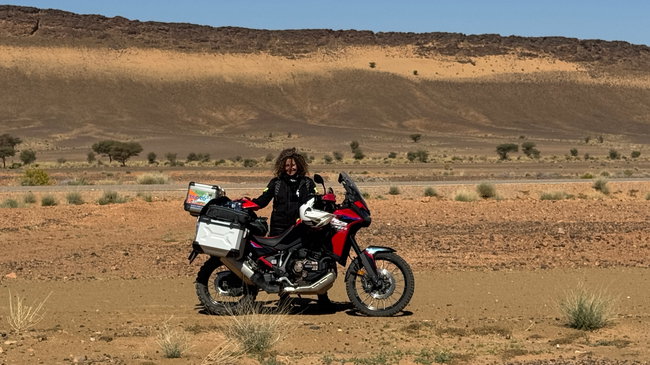Voyage moto au Maroc MM8 – Les premières dunes sur la route du Sahara Paysage aride sur la route du Sahara, avec du sable au bord de la chaussée et les premières petites dunes à l'horizon